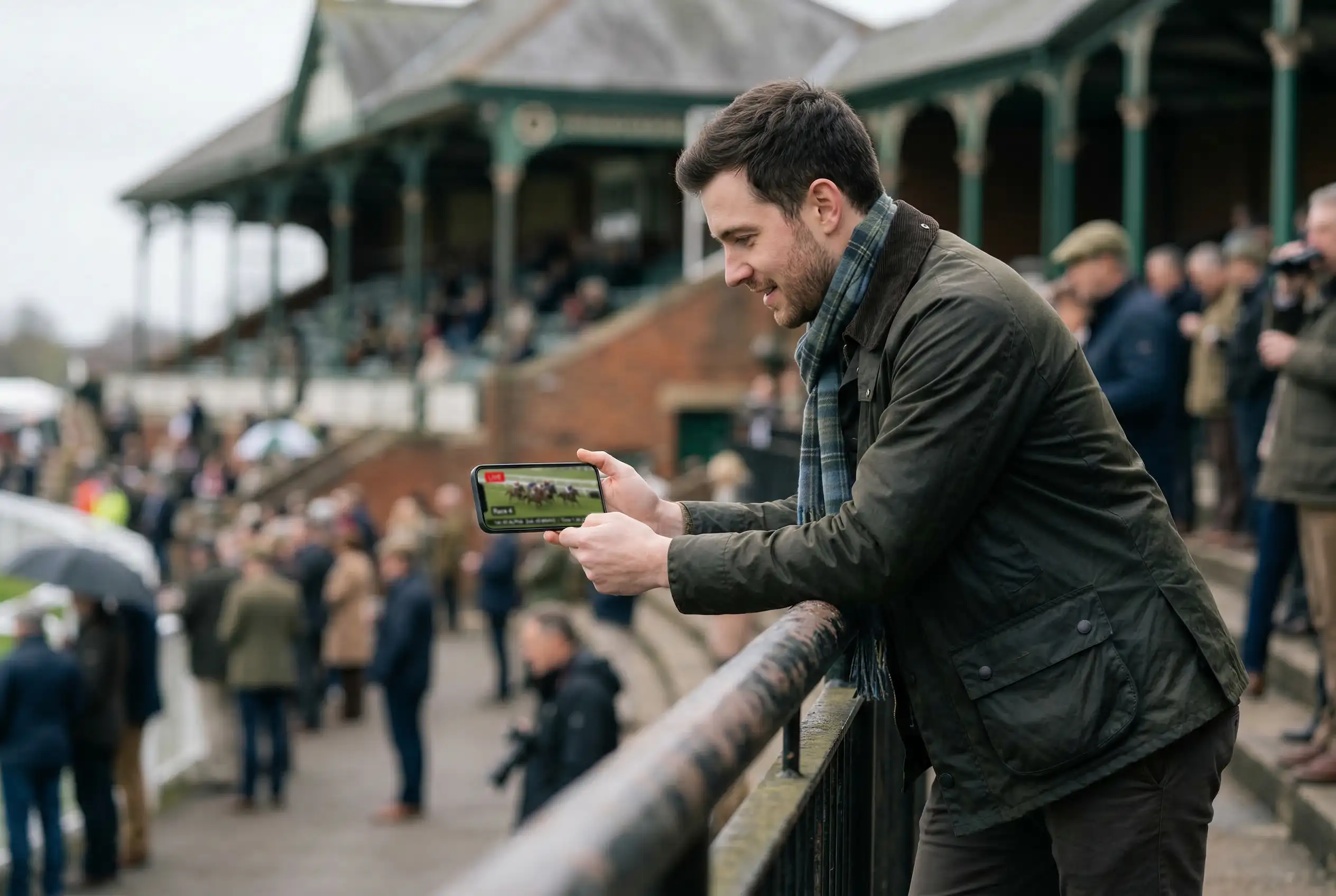 Punter watching live horse racing on a smartphone at a UK racecourse with the track visible in the background